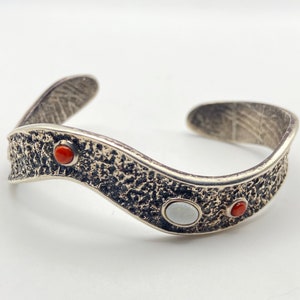 May include: A silver-toned cuff bracelet with a textured surface and a wavy design. The bracelet features three inlaid gemstones: two red and one white, adding a pop of color to the jewelry.