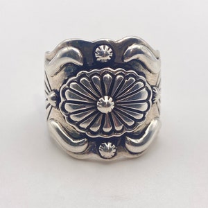 May include: A silver ring with a floral design. The ring is wide and has a raised center with a flower design. The flower has a center circle and petals that radiate outwards.