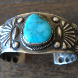 K&ouml;nnte beinhalten: Ein silbernes Armreif mit einem T&uuml;rkis-Stein in der Mitte. Das Armband ist mit silbernen Perlen und geometrischen Mustern verziert.