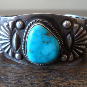 K&ouml;nnte beinhalten: Ein silbernes Armreif mit einem T&uuml;rkis-Stein in der Mitte. Das Armband ist mit silbernen Perlen und einem Seil-Design verziert.