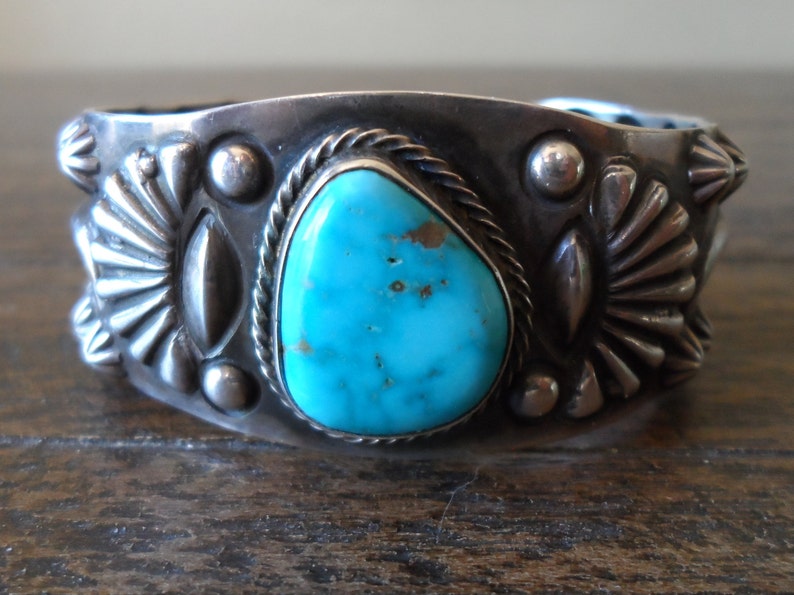 K&ouml;nnte beinhalten: Ein silbernes Armreif mit einem T&uuml;rkis-Stein in der Mitte. Das Armband ist mit silbernen Perlen und einem Seil-Design verziert.