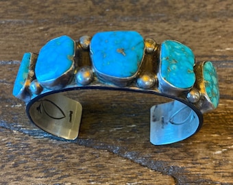 May include: A silver cuff bracelet featuring five turquoise stones arranged in a row. The stones are a vibrant blue-green colour and have a slightly irregular shape. The bracelet is stamped with a small leaf design on the back.
