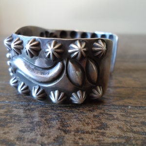 K&ouml;nnte beinhalten: Ein silbernes Armreif mit einem dekorativen Design aus stilisierten Bl&auml;ttern und Blumen. Das Armband ist mit einer Reihe von sternf&ouml;rmigen Akzenten verziert.
