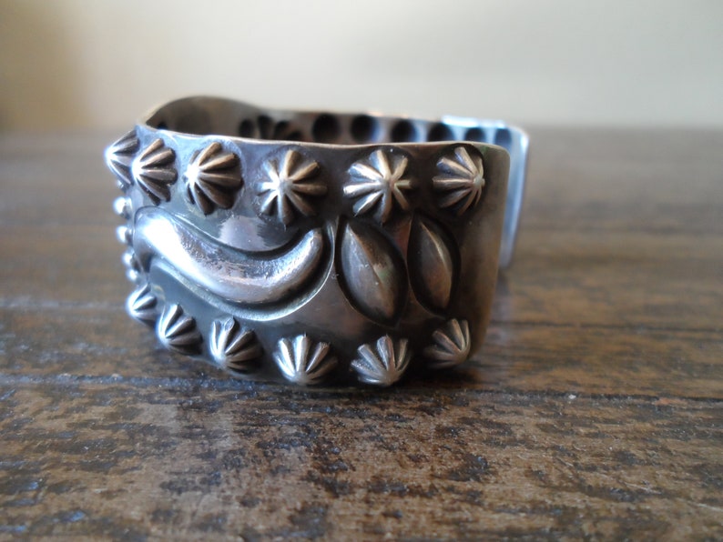 K&ouml;nnte beinhalten: Ein silbernes Armreif mit einem dekorativen Design aus stilisierten Bl&auml;ttern und Blumen. Das Armband ist mit einer Reihe von sternf&ouml;rmigen Akzenten verziert.