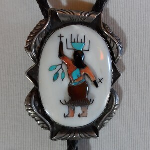 May include: Oval bolo tie with a white inlaid center featuring a Native American figure with turquoise, orange, and brown accents. The figure is set in a silver-toned frame with a decorative leaf design. The tie has a black leather cord.