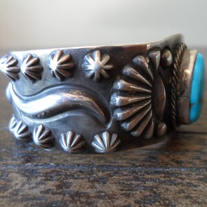 K&ouml;nnte beinhalten: Ein silbernes Armreif mit einem T&uuml;rkis-Stein in der Mitte. Das Armband ist mit einem Wellendesign und Sternburst-Mustern verziert.