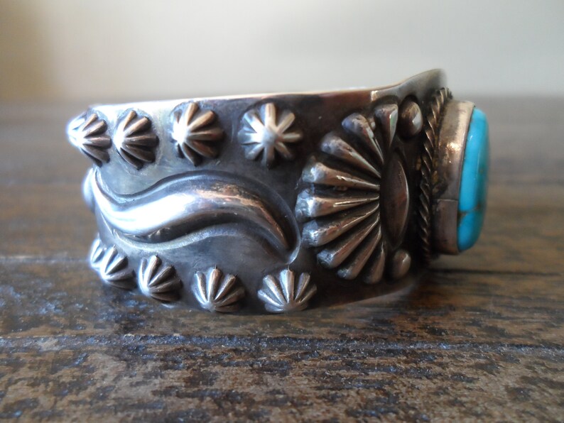 K&ouml;nnte beinhalten: Ein silbernes Armreif mit einem T&uuml;rkis-Stein in der Mitte. Das Armband ist mit einem Wellendesign und Sternburst-Mustern verziert.