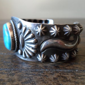 K&ouml;nnte beinhalten: Ein silbernes Armreif mit einem T&uuml;rkis-Stein in der Mitte. Das Armband ist mit einem sich wiederholenden Muster aus Sternchen und einer Wellenlinie verziert.