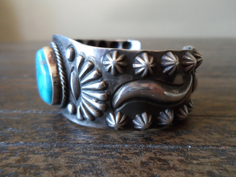 K&ouml;nnte beinhalten: Ein silbernes Armreif mit einem T&uuml;rkis-Stein in der Mitte. Das Armband ist mit einem sich wiederholenden Muster aus Sternchen und einer Wellenlinie verziert.