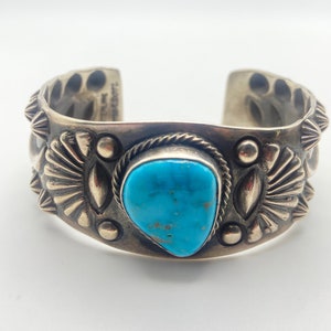K&ouml;nnte beinhalten: Ein silbernes Armreif mit einem T&uuml;rkis-Stein in der Mitte. Das Armband ist mit komplizierten Silberdesigns verziert.