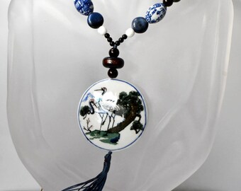 May include: A blue and white porcelain pendant necklace with a tassel. The pendant features a painted design of two cranes standing on a branch in a landscape. The necklace is made with blue and white porcelain beads, brown wooden beads, and black beads.