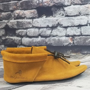 Puede incluir: Un par de mocasines de ante amarillo mostaza con un lazo de cuero negro y una suela negra. Los mocasines tienen un logotipo de "Red Jacket Trading Co." en el lateral.