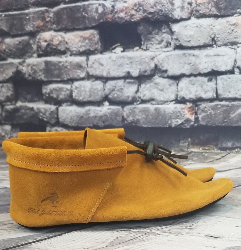 Puede incluir: Un par de mocasines de ante amarillo mostaza con un lazo de cuero negro y una suela negra. Los mocasines tienen un logotipo de "Red Jacket Trading Co." en el lateral.