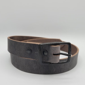 Puede incluir: Un cinturón de cuero gris oscuro con una hebilla de metal negro. El cinturón está enrollado y la hebilla es visible.