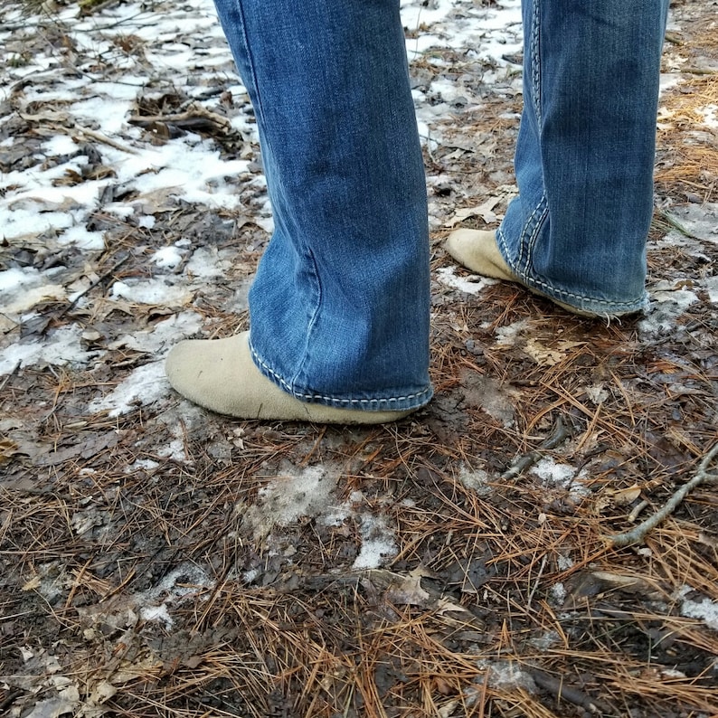 Puede incluir: Un par de pantalones vaqueros azules con los dobladillos inferiores enrollados, mostrando los zapatos de gamuza marr&oacute;n claro. Los zapatos tienen una punta puntiaguda y un tac&oacute;n bajo. La persona est&aacute; de pie sobre un lecho de agujas de pino marrones y nieve.
