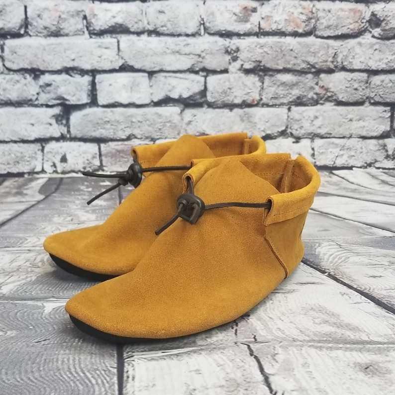 Puede incluir: Un par de mocasines de ante amarillo mostaza con cordones de cuero marr&oacute;n. Los mocasines tienen una punta puntiaguda y un tac&oacute;n bajo.
