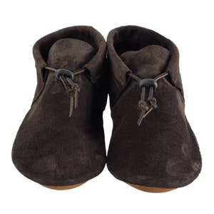 Puede incluir: Un par de mocasines de ante marr&oacute;n con cordones de cuero y un cierre de bot&oacute;n negro.
