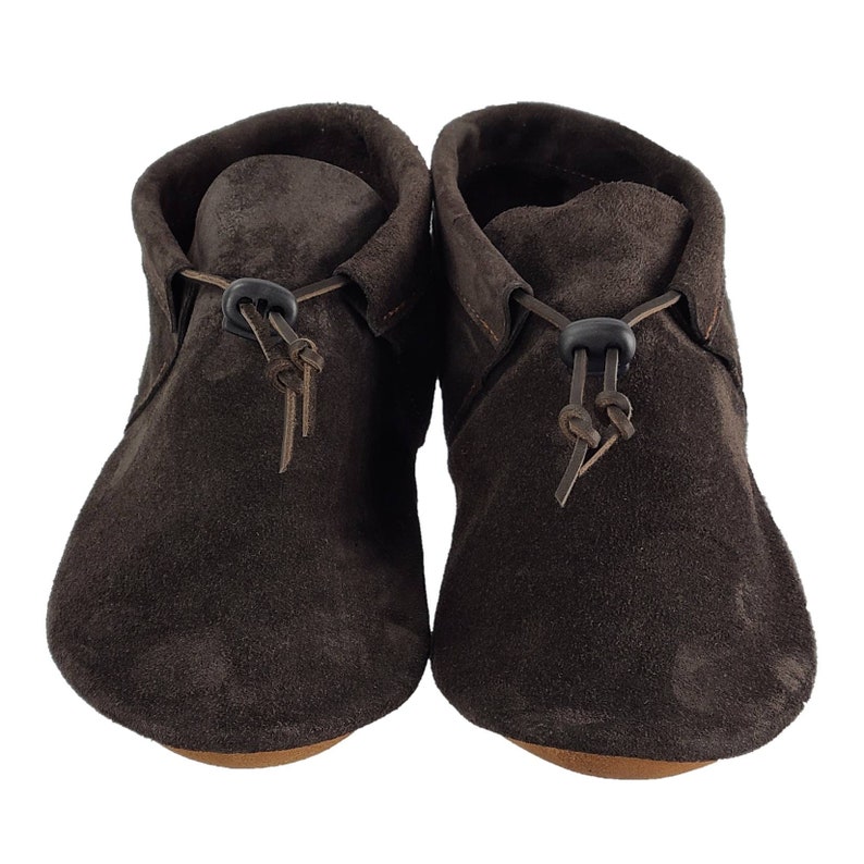 Puede incluir: Un par de mocasines de ante marr&oacute;n con cordones de cuero y un cierre de bot&oacute;n negro.