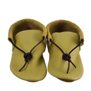 May include: A pair of light green leather moccasins with brown leather laces and black buckles. The moccasins have a soft, suede-like texture and a simple, minimalist design.