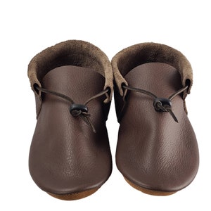 May include: A pair of brown leather moccasins with a fold-over cuff and a tie closure. The moccasins have a textured leather surface and a rounded toe.