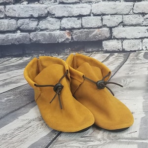Puede incluir: Un par de botines de tobillo de ante amarillo mostaza con cordones marrones y hebillas negras.