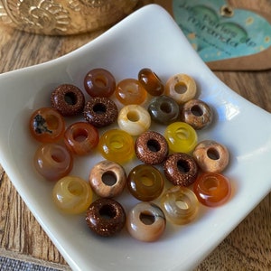 Może przedstawiać: Biała ceramiczna miseczka wypełniona różnymi małymi, okrągłymi koralikami w kształcie pączka. Korale są wykonane z naturalnego kamienia i występują w odcieniach brązu, pomarańczy, żółci i beżu.