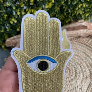 Peut inclure: Écusson brodé en forme de main de Hamsa dorée avec un motif d'œil bleu et noir. Le patch a une bordure blanche et est probablement destiné à des fins décoratives. Le motif est détaillé avec des coutures visibles, et le patch est probablement en tissu.