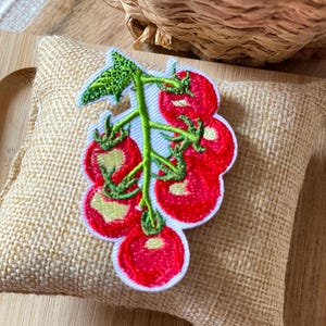 Può includere: Toppa ricamata raffigurante un grappolo di pomodori rossi con steli e foglie verdi. La toppa è bordata di bianco ed è posta su uno sfondo di tela di iuta beige. I pomodori hanno un colore rosso vivo.