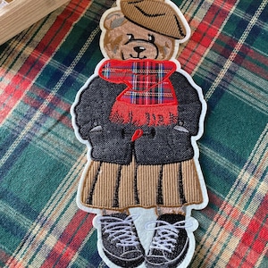 Peut inclure: Écusson brodé d'un ours en peluche portant un béret marron, une écharpe à carreaux rouge, un manteau noir, une jupe plissée et des baskets noires à lacets blancs. L'ours est sur un fond à carreaux vert et rouge.