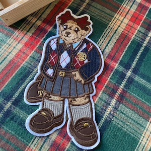 Könnte beinhalten: Gestickter Aufnäher mit einem Teddybären in Schuluniform. Der Bär trägt einen marineblauen Blazer mit rotem und weißem Argyle-Muster, einen Faltenrock und braune Loafer. Der Aufnäher hat einen weißen Rand.
