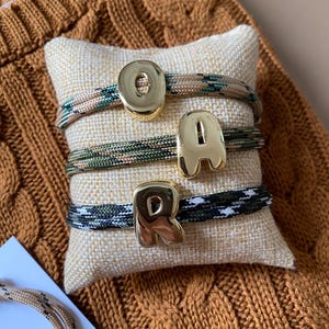 Peut inclure: Trois bracelets en corde avec des breloques dorées formant le mot "OAR" sont présentés sur un petit coussin beige. Les bracelets présentent des motifs camouflage et noir et blanc. L'arrière-plan est un pull marron.