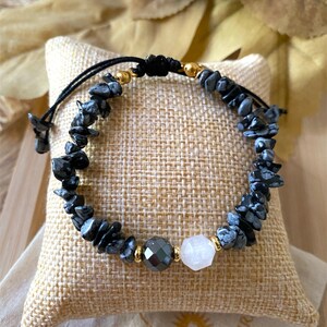 Peut inclure: Un bracelet en pierres noires et blanches avec un cordon noir. Le bracelet comporte une perle grise facettée et une perle blanche hexagonale, avec des accents dorés. Le bracelet est présenté sur un coussin beige.