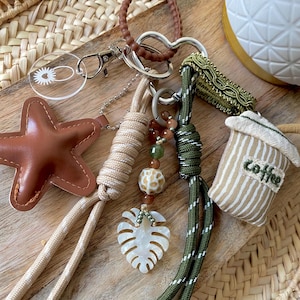 Puede incluir: Una colección de llaveros y colgantes con varios diseños. Incluye una estrella de cuero marrón, un colgante circular transparente, un colgante de taza de café y un colgante de hoja de monstera. Los llaveros presentan detalles de cuerda y cuentas en tonos neutros y verdes.