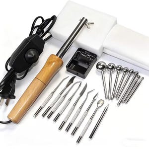 May include: A soldering iron with a wooden handle, black power cord, and a selection of metal tools. The tools include various blade shapes and ball-tipped tools, a black metal stand, and a white foam block.