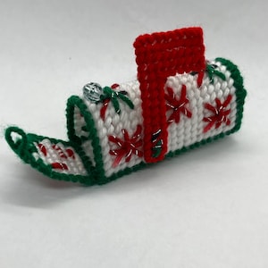 May include: A handmade, miniature mailbox ornament. The white body is decorated with red and silver starbursts and green accents. The flag and trim are red and green. A small, clear bead is attached to the top.