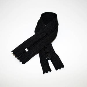 Puede incluir: Cremallera negra sobre fondo blanco. La cremallera está cerrada, con el tirador en su lugar. Los dientes de la cremallera son visibles y la cinta de tela es de color negro liso. Adecuada para diversos proyectos de costura.