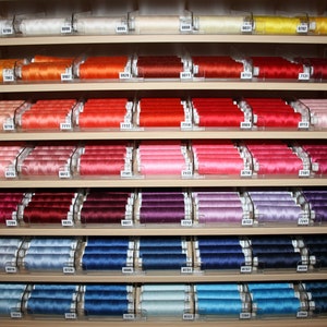 May include: A shelf display of spools of thread in various colours, including white, orange, red, pink, purple, blue, and light blue. Each spool has a number printed on it.