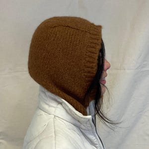 May include: A brown knitted hooded scarf or neck warmer. The scarf is made of a soft, fuzzy material and is worn by a person with long dark hair.