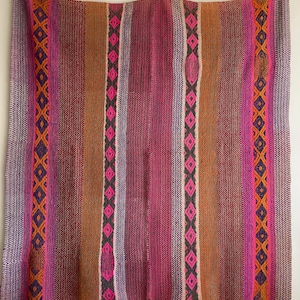 May include: A woven throw blanket with a colourful striped pattern in shades of pink, orange, purple, and brown. The blanket has a geometric design with diamond shapes.