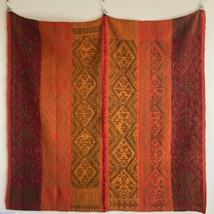 May include: A woven tapestry featuring a geometric pattern in shades of orange, red, and brown. The tapestry is made of wool and has a fringed edge.