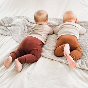 Pode incluir: Dois beb&ecirc;s vestindo leggings marrons e camisas cinzas est&atilde;o deitados de bru&ccedil;os em um cobertor branco.