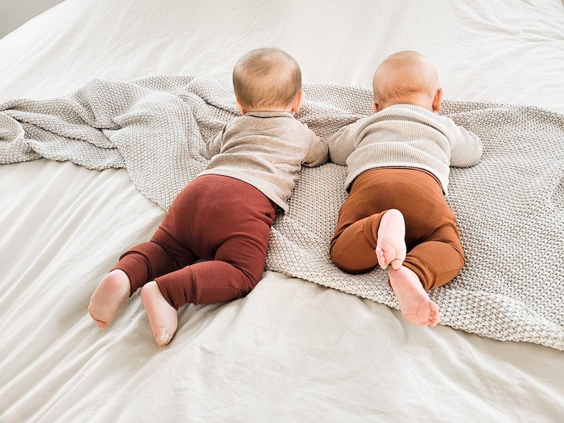 Pode incluir: Dois beb&ecirc;s vestindo leggings marrons e camisas cinzas est&atilde;o deitados de bru&ccedil;os em um cobertor branco.