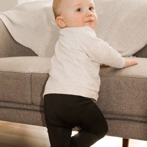 Pode incluir: Um beb&ecirc; com uma camisa branca de manga comprida e leggings pretos est&aacute; em p&eacute; em um sof&aacute; cinza. O beb&ecirc; est&aacute; olhando por cima do ombro para a c&acirc;mera.