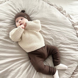 Pode incluir: Um beb&ecirc; vestindo um chap&eacute;u de tric&ocirc; marrom com um n&oacute; no topo e um su&eacute;ter creme com cal&ccedil;as marrons. O beb&ecirc; est&aacute; deitado em um cobertor marrom claro com babados.