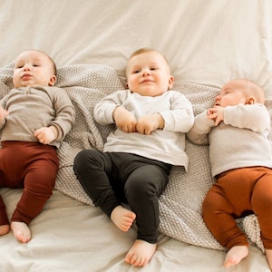 Pode incluir: Tr&ecirc;s beb&eacute;s deitados em um cobertor branco com um cobertor de malha cinza. O beb&ecirc; &agrave; esquerda est&aacute; vestindo uma camisa marrom e cal&ccedil;as marrons. O beb&ecirc; do meio est&aacute; vestindo uma camisa branca e cal&ccedil;as pretas. O beb&ecirc; &agrave; direita est&aacute; vestindo uma camisa branca e cal&ccedil;as marrons.