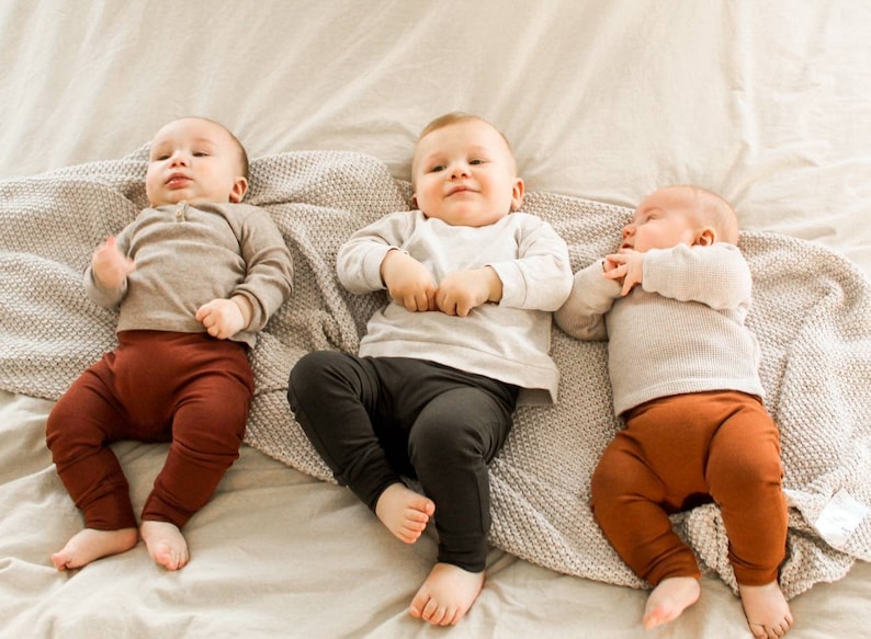Pode incluir: Tr&ecirc;s beb&eacute;s deitados em um cobertor branco com um cobertor de malha cinza. O beb&ecirc; &agrave; esquerda est&aacute; vestindo uma camisa marrom e cal&ccedil;as marrons. O beb&ecirc; do meio est&aacute; vestindo uma camisa branca e cal&ccedil;as pretas. O beb&ecirc; &agrave; direita est&aacute; vestindo uma camisa branca e cal&ccedil;as marrons.