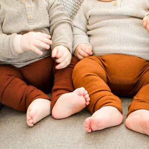 Pode incluir: Dois beb&ecirc;s vestindo cal&ccedil;as marrons e camisas cinzas de manga comprida est&atilde;o sentados em um sof&aacute; cinza.