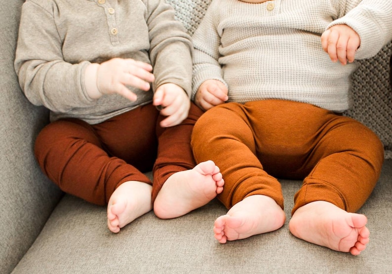 Pode incluir: Dois beb&ecirc;s vestindo cal&ccedil;as marrons e camisas cinzas de manga comprida est&atilde;o sentados em um sof&aacute; cinza.