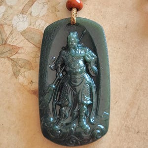 May include: A dark green jade pendant necklace with a carved warrior figure. The rectangular pendant with rounded corners hangs from a gold-coloured cord. The necklace includes a decorative element with black, red, and blue beads.
