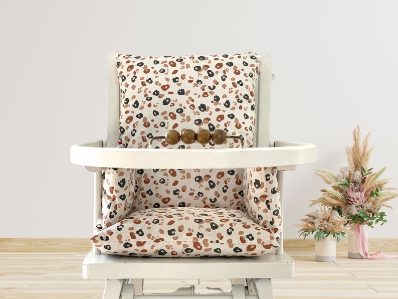 Op de afbeelding: Witte hoge stoel met een kussen met luipaardprint en een houten dienblad. Het kussen heeft een bruin, zwart en wit patroon.